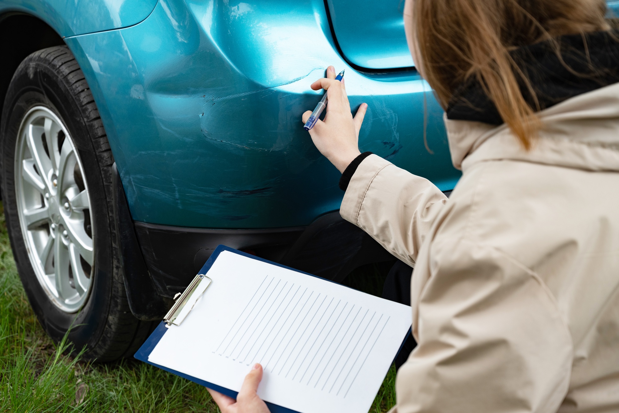 Person inspecting car bumper damage with clipboard outdoors.